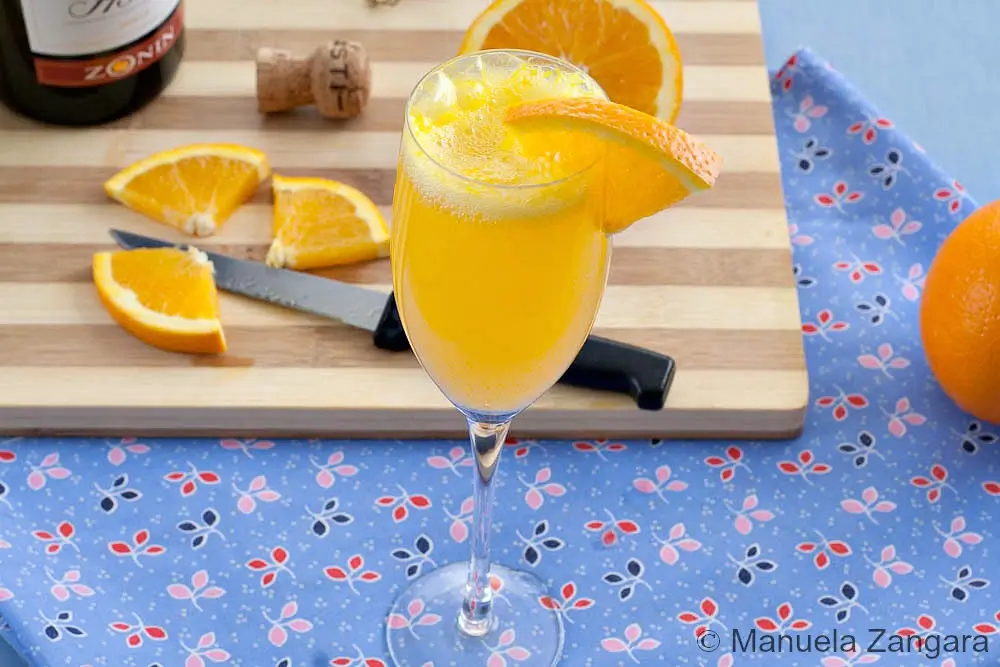 Mimosa cocktail in a flute on a board with sliced oranges and a cork beside it.
