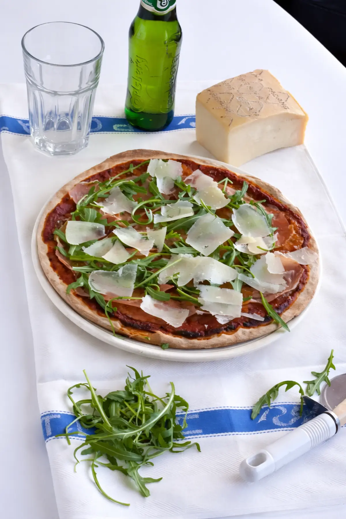 Pizza topped with prosciutto, fresh rocket, and shaved cheese on a white table setting.