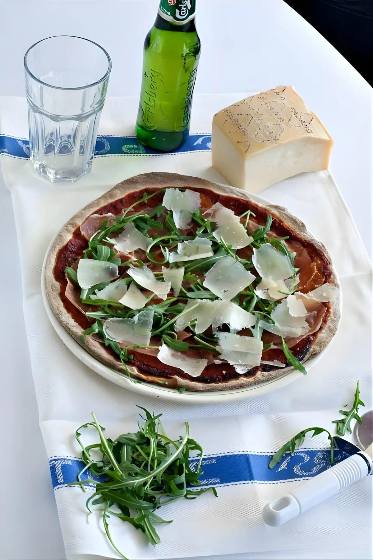 Pizza topped with prosciutto, rocket, and Grana slivers served with cheese and a drink on the side.