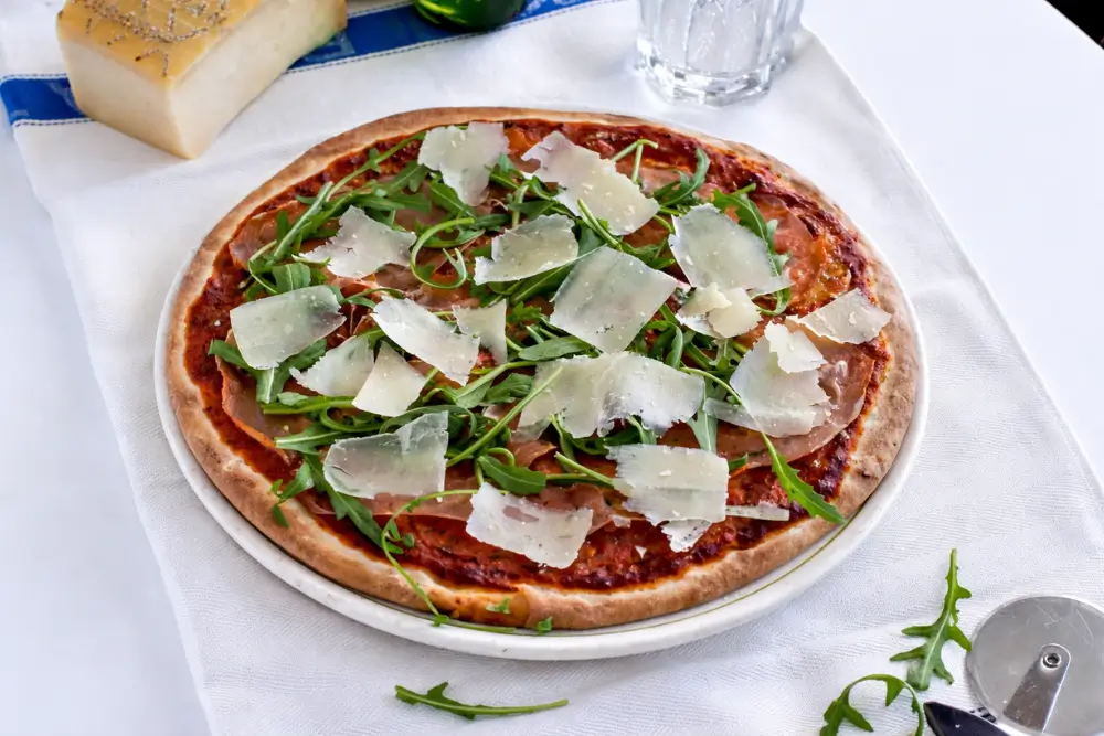 Pizza with prosciutto, fresh rocket, and shaved cheese served on a plate with beer and Parmigiano.