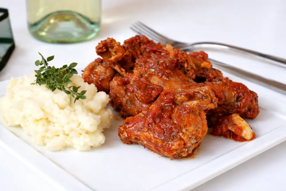 Braised rabbit pieces covered in tomato sauce served beside creamy mashed potatoes with herbs.