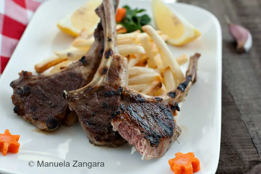Grilled lamb cutlets served with fries and lemon wedges on a white plate.