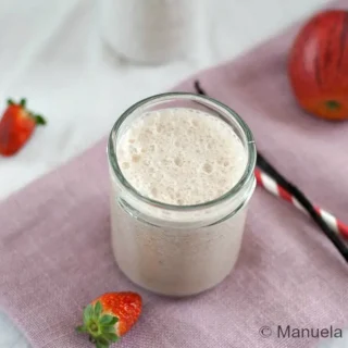 Close-up of a blended fruit smoothie with apple and strawberries in the background.