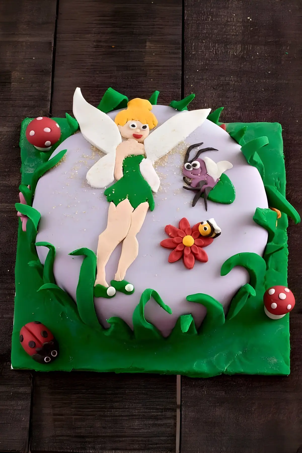 Tinkerbell fondant cake decorated with flowers, grass, Blaze the firefly, a bee, and toadstools on a lavender and green base.