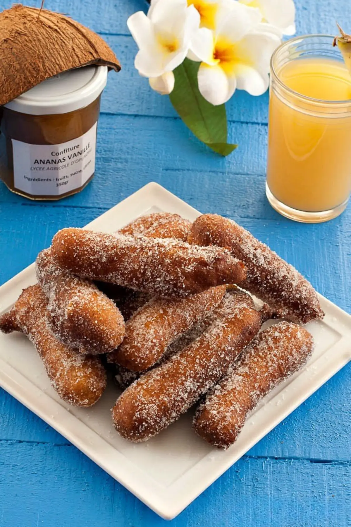 A plate of sugared Firi Firi served with pineapple jam, coconut and juice on a blue table.