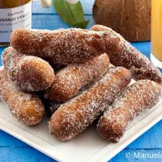 A stack of sugared Firi Firi on a white plate with coconut and juice in the background.