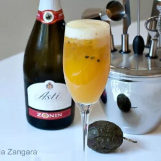 A tall Champagne flute filled with mango and passion fruit cocktail topped with foam, next to a bottle of Asti and bar tools.