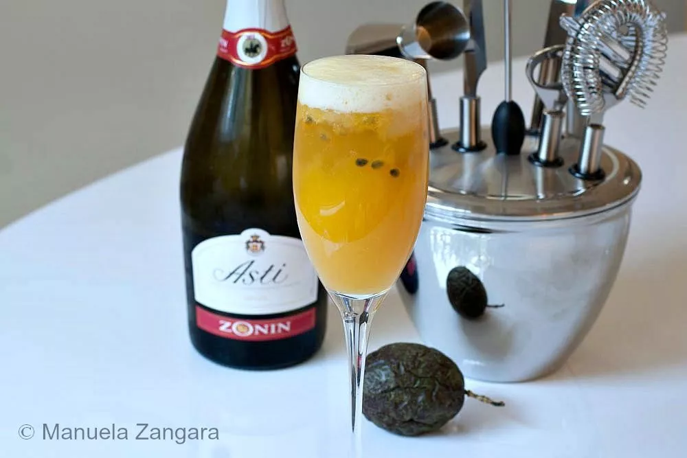 A tall Champagne flute filled with mango and passion fruit cocktail topped with foam, next to a bottle of Asti and bar tools.