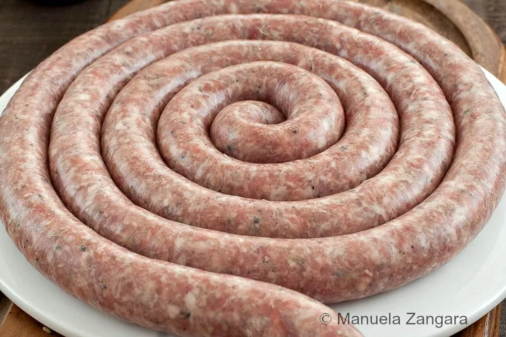 Traditional Sicilian fennel sausage coiled on a white plate with fennel seeds on the side.