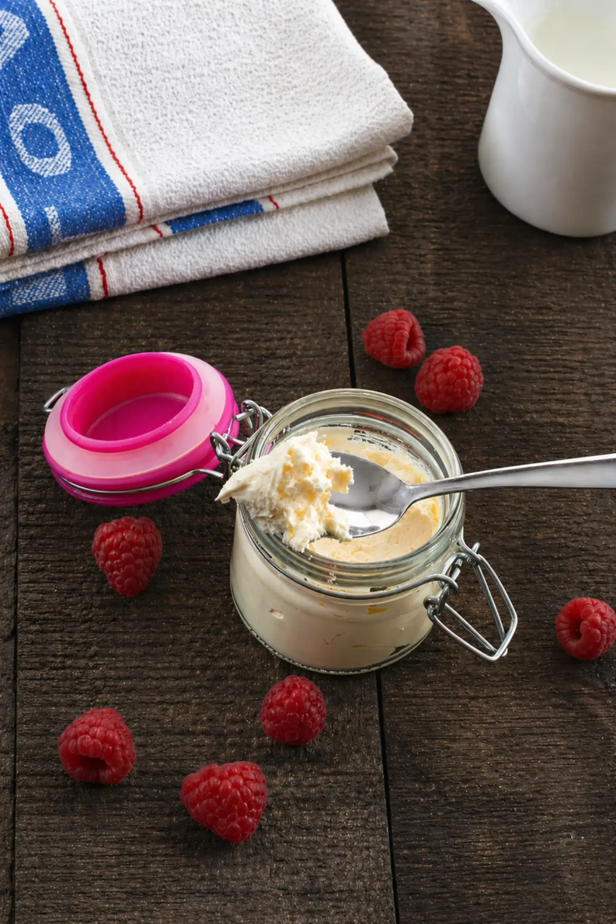 Spoon scooping creamy Italian cheese spread from a glass jar surrounded by raspberries.