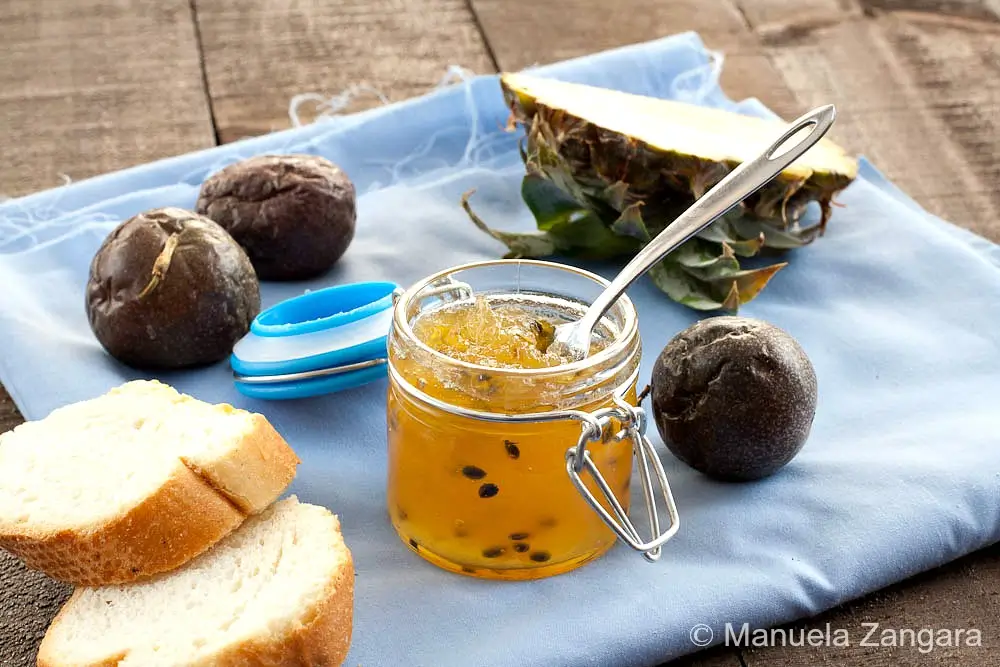 Jar of pineapple and passion fruit jam surrounded by sliced bread, passion fruit, and fresh pineapple.