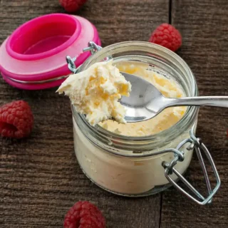 Spoon lifting homemade mascarpone from a glass jar with raspberries on a wooden table.
