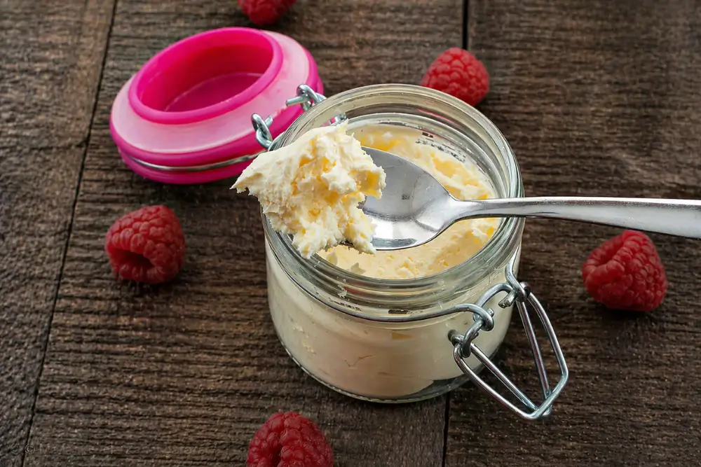 Spoon lifting homemade mascarpone from a glass jar with raspberries on a wooden table.