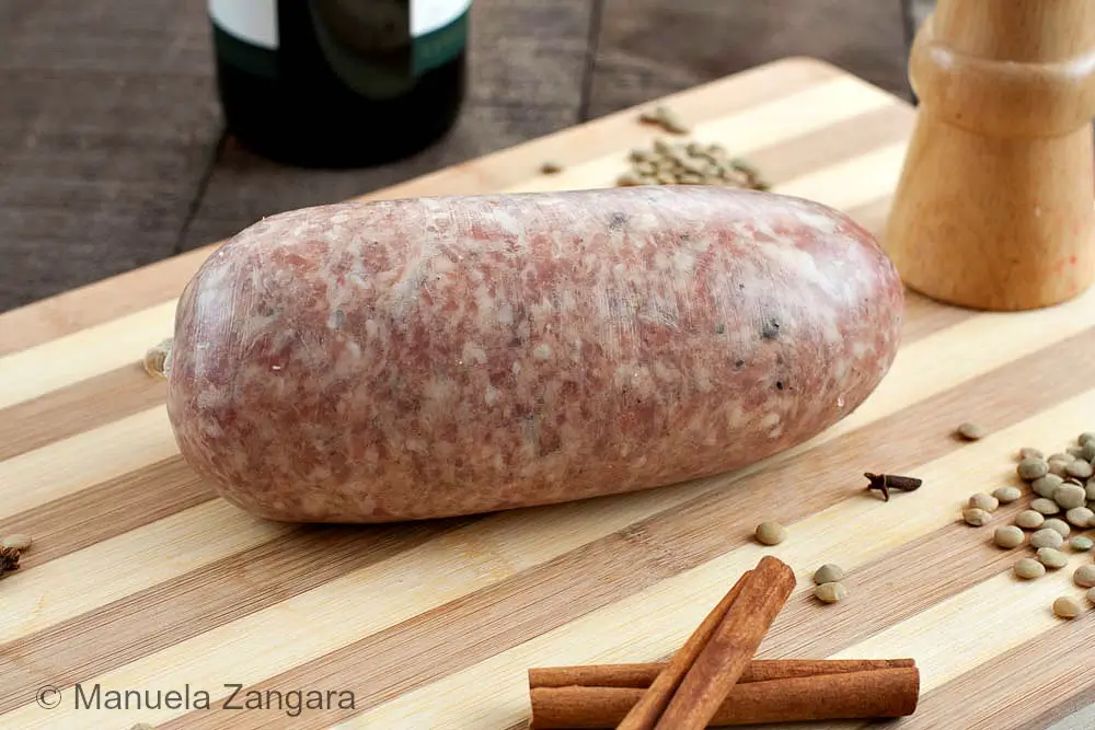 A whole uncooked cotechino resting on a striped chopping board with lentils, spices, and a wooden pepper mill.