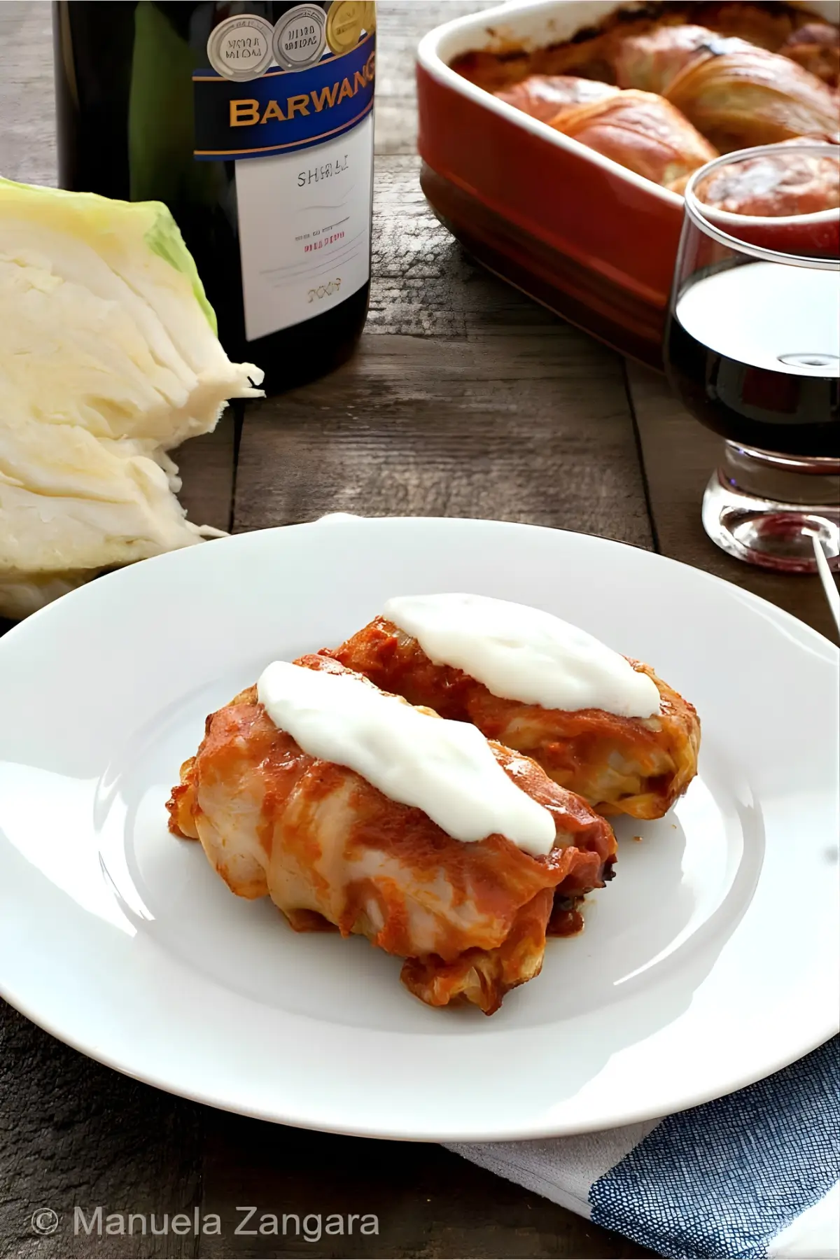 Two cabbage rolls served on a white plate with sour cream, wine, and baked rolls in the background.