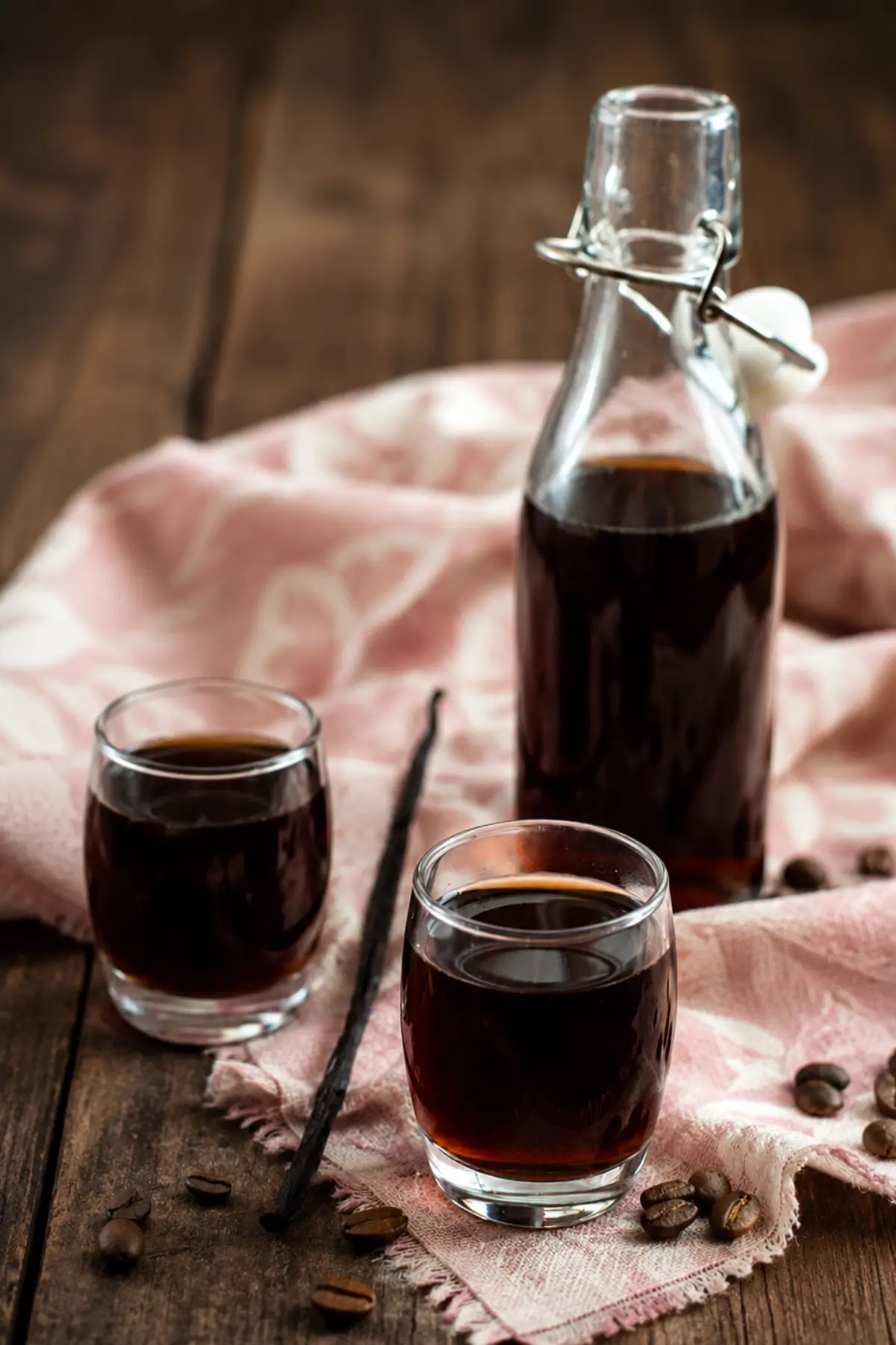 Two small glasses of Coffee and Vanilla Liqueur beside a glass bottle with coffee beans and vanilla pods on a wooden table.