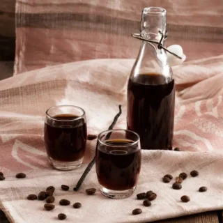 Homemade coffee liqueur served in small glasses with a swing-top bottle, coffee beans, and vanilla on a soft cloth background.