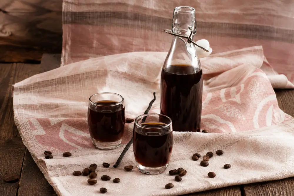 Homemade coffee liqueur served in small glasses with a swing-top bottle, coffee beans, and vanilla on a soft cloth background.
