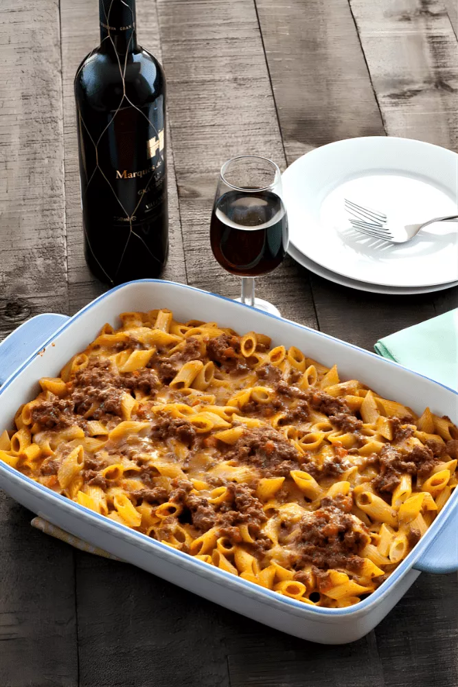 Traditional Italian Pasta al Forno in a white baking dish with a bottle of red wine and a serving plate beside it.