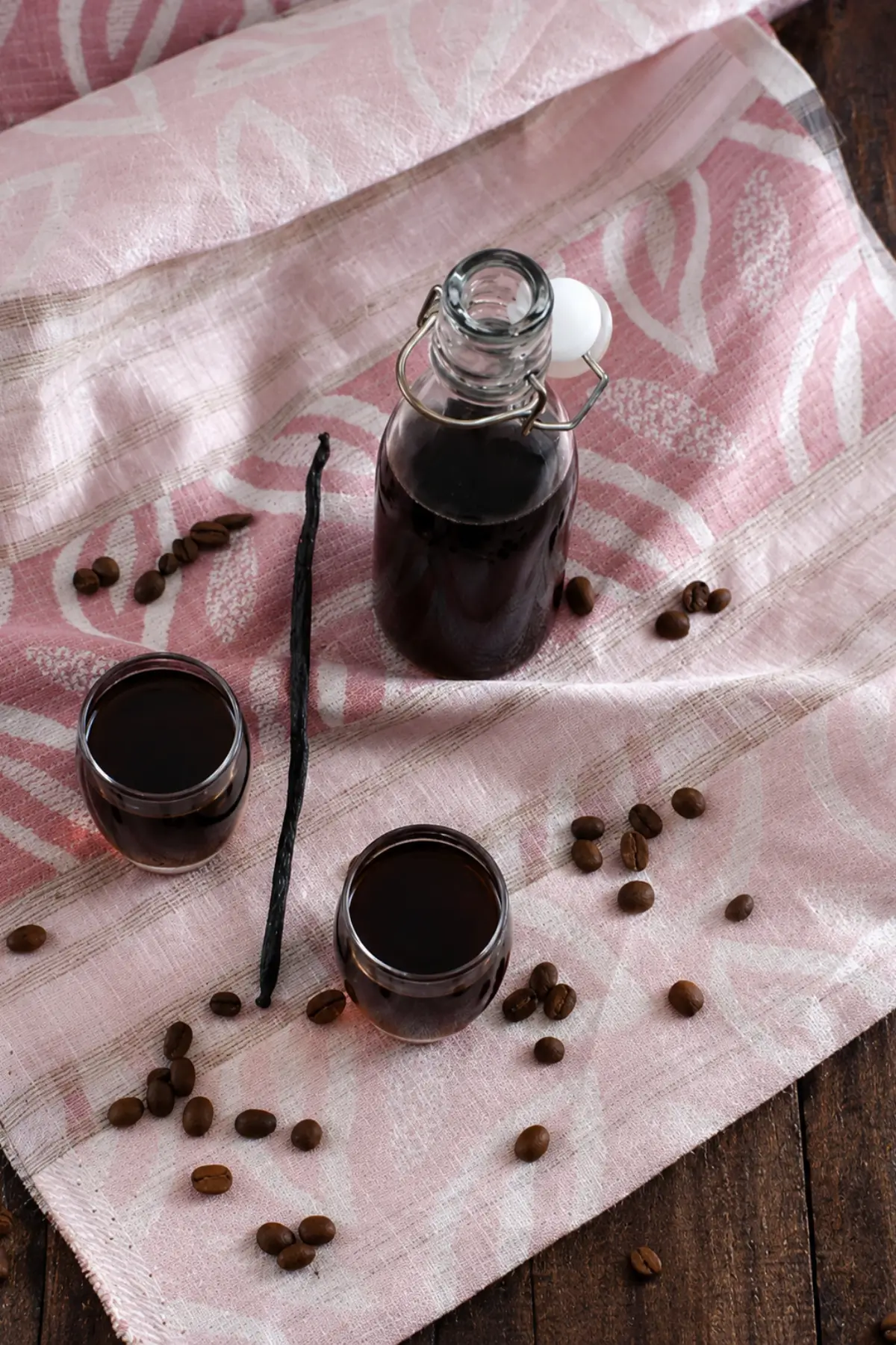 Bottle and glasses of vanilla coffee liqueur served with whole coffee beans and a vanilla pod on a soft cloth background.