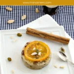 Couscous pudding topped with a candied orange slice, pistachios, and almonds with light syrup on the plate.