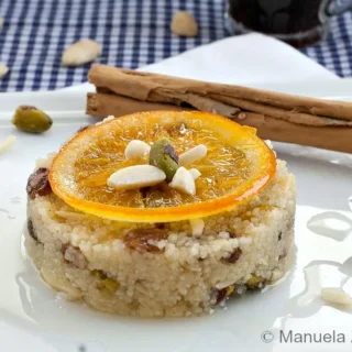 Couscous pudding topped with a candied orange slice, pistachios, and almonds with light syrup on the plate.