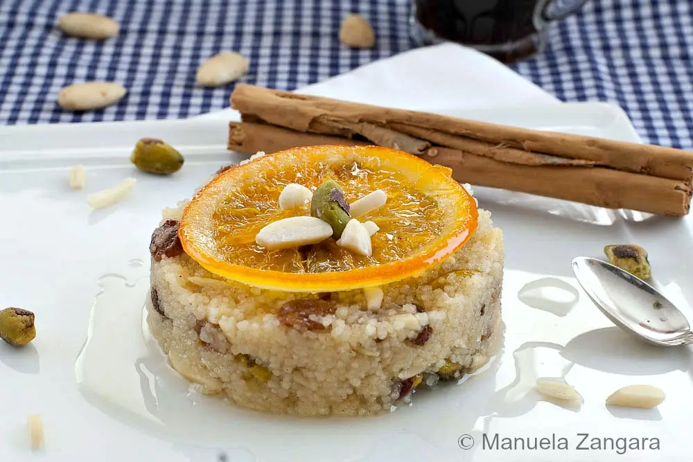 Couscous pudding topped with a candied orange slice, pistachios, and almonds with light syrup on the plate.