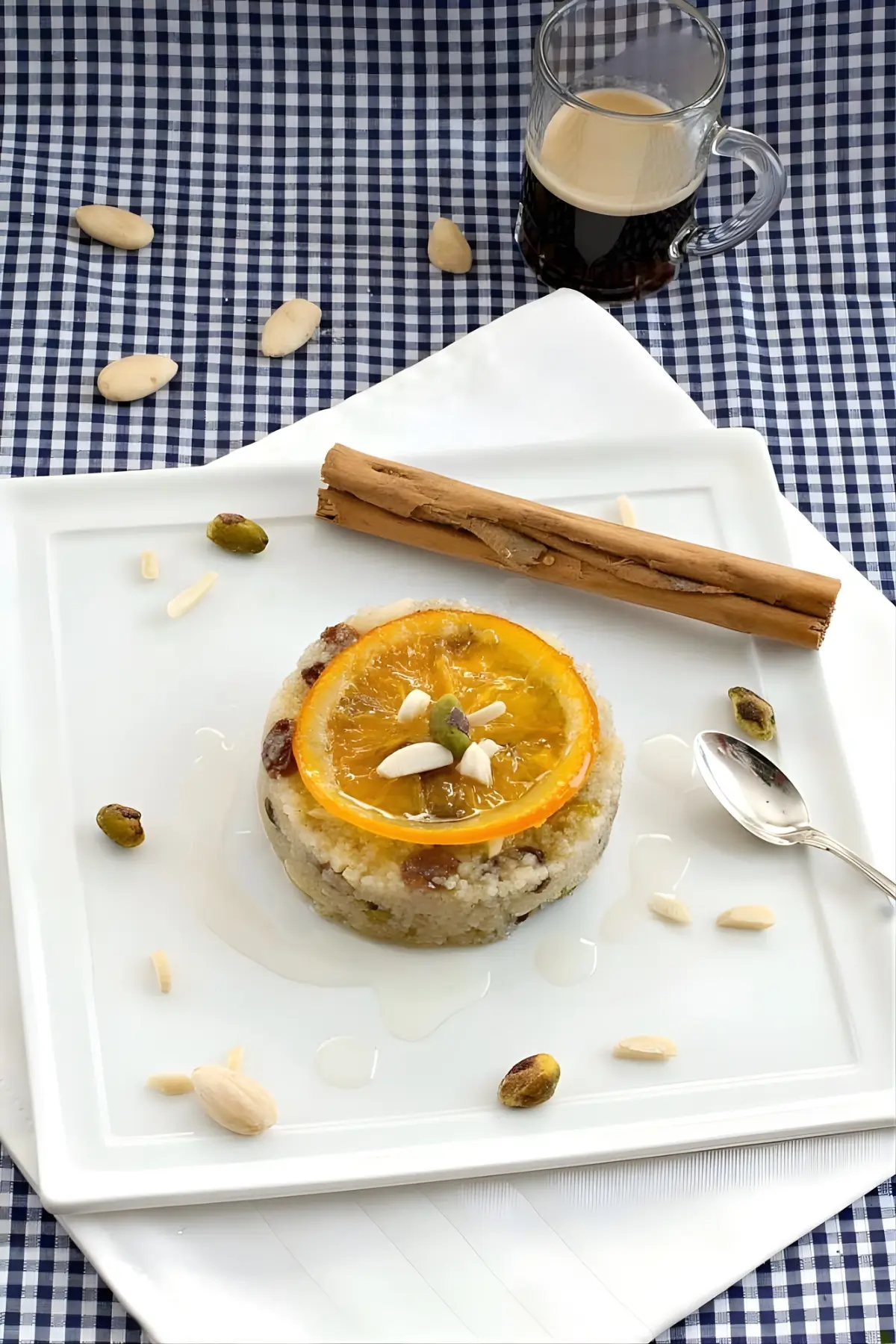 A shaped couscous pudding topped with a candied orange slice, nuts, and syrup on a square white plate with cinnamon sticks.