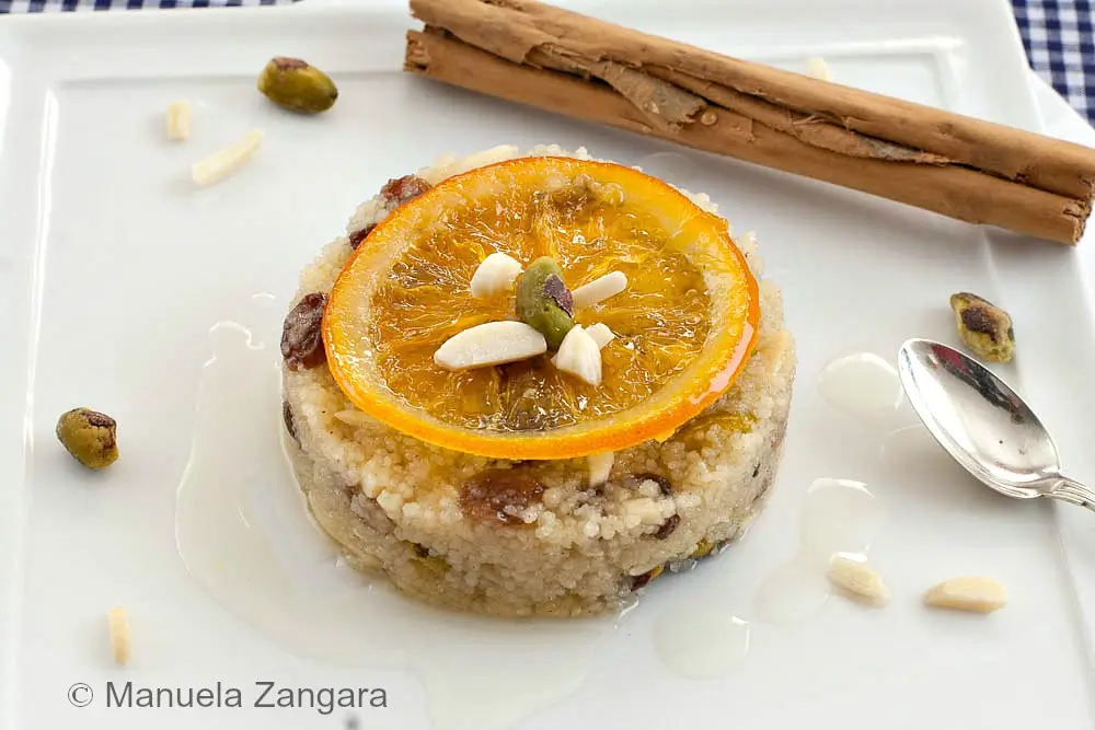 North African couscous pudding topped with a candied orange slice, nuts and syrup on a white plate.