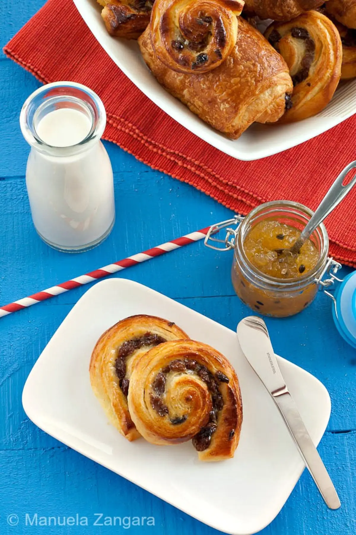 Two baked pain aux raisins on a white plate with milk and jam on the side.