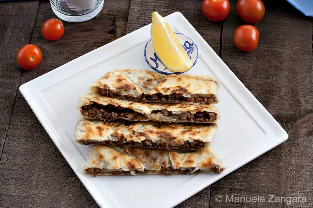 Sliced stuffed flatbread with browned spots, served on a white plate with a lemon wedge and cherry tomatoes.