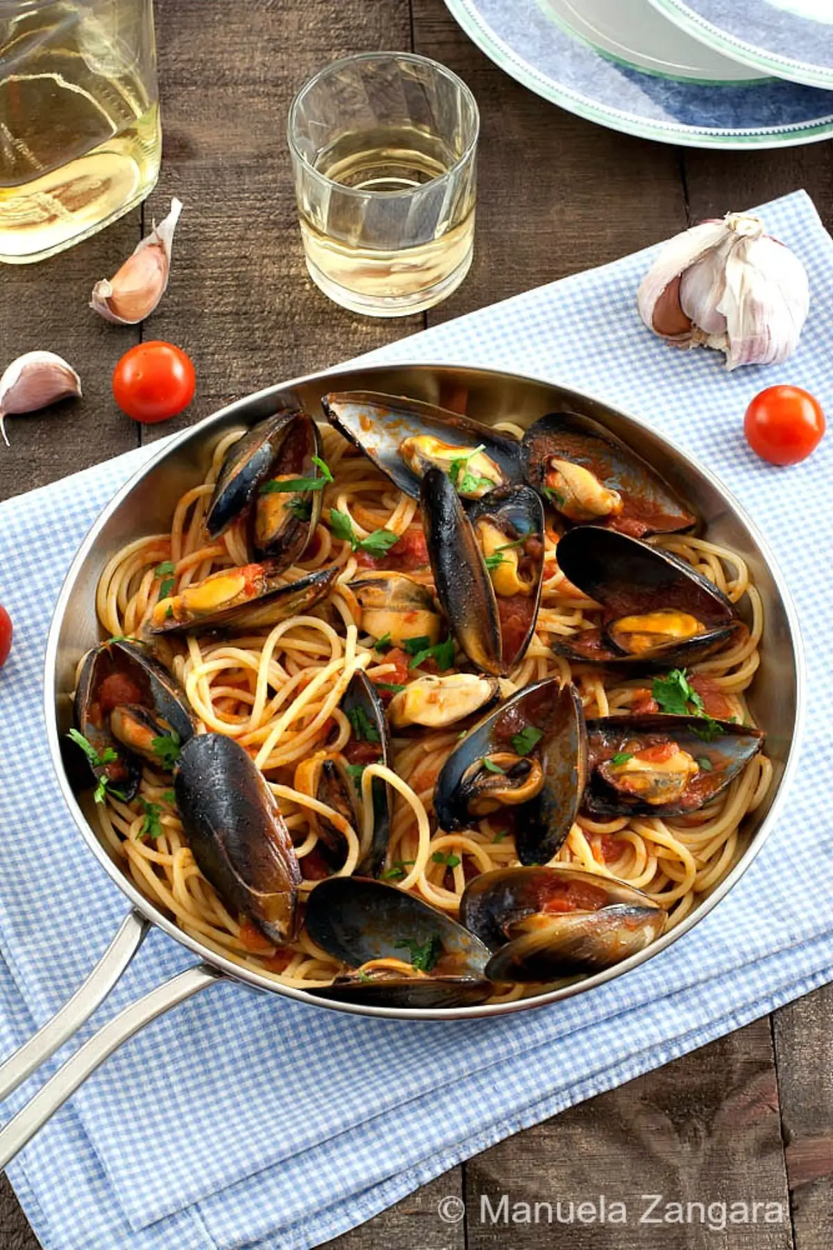 Spaghetti with mussels, cherry tomatoes, and parsley served in a pan with a glass of white wine.