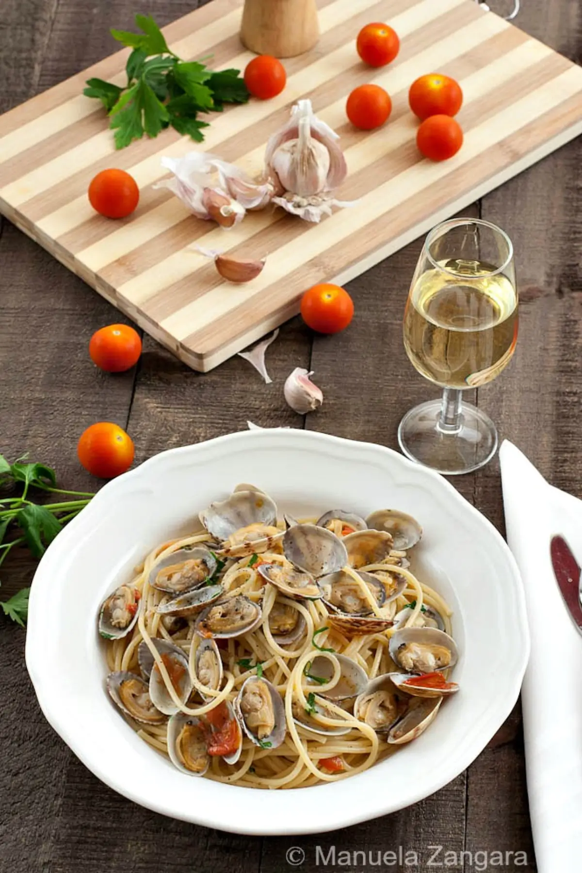 Spaghetti with clams, cherry tomatoes, and parsley served with a glass of white wine on a wooden table.