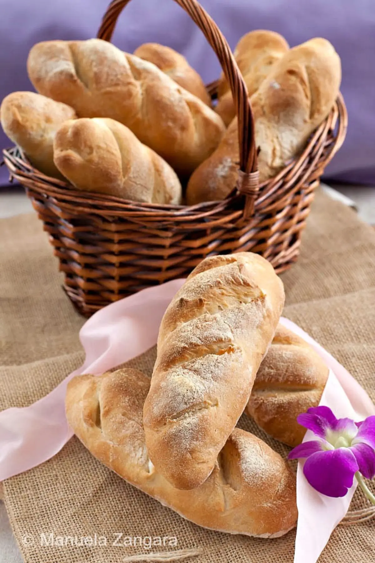 Freshly baked Italian bread rolls arranged on burlap beside a basket.