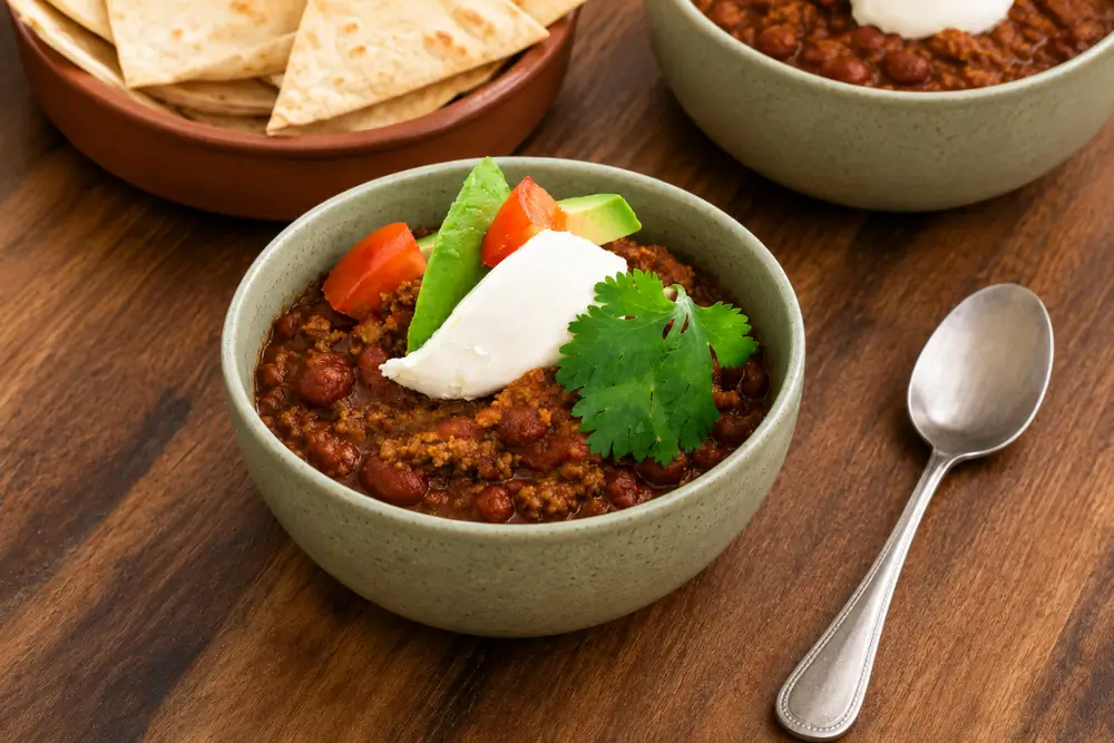 Bowl of chilli with beans topped with avocado, tomato, sour cream and fresh coriander, with tortilla chips alongside.