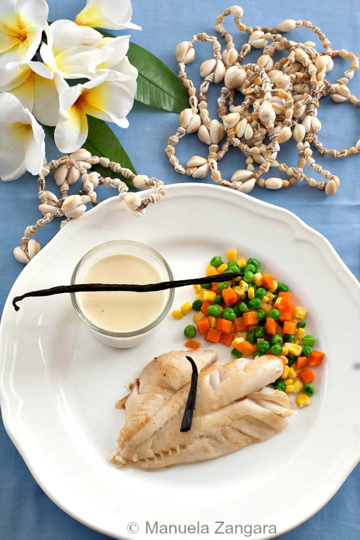 Overhead shot of fish fillet, vanilla sauce, and vegetables arranged on a plate with island décor.