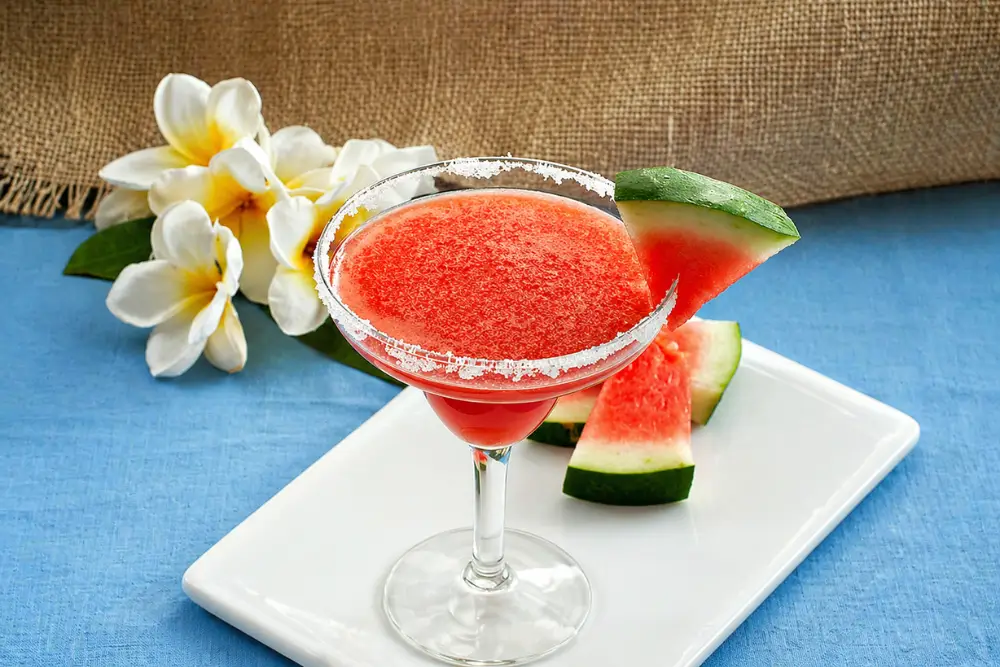 Watermelon margarita in a sugar-rimmed glass with fresh watermelon slices on the side.
