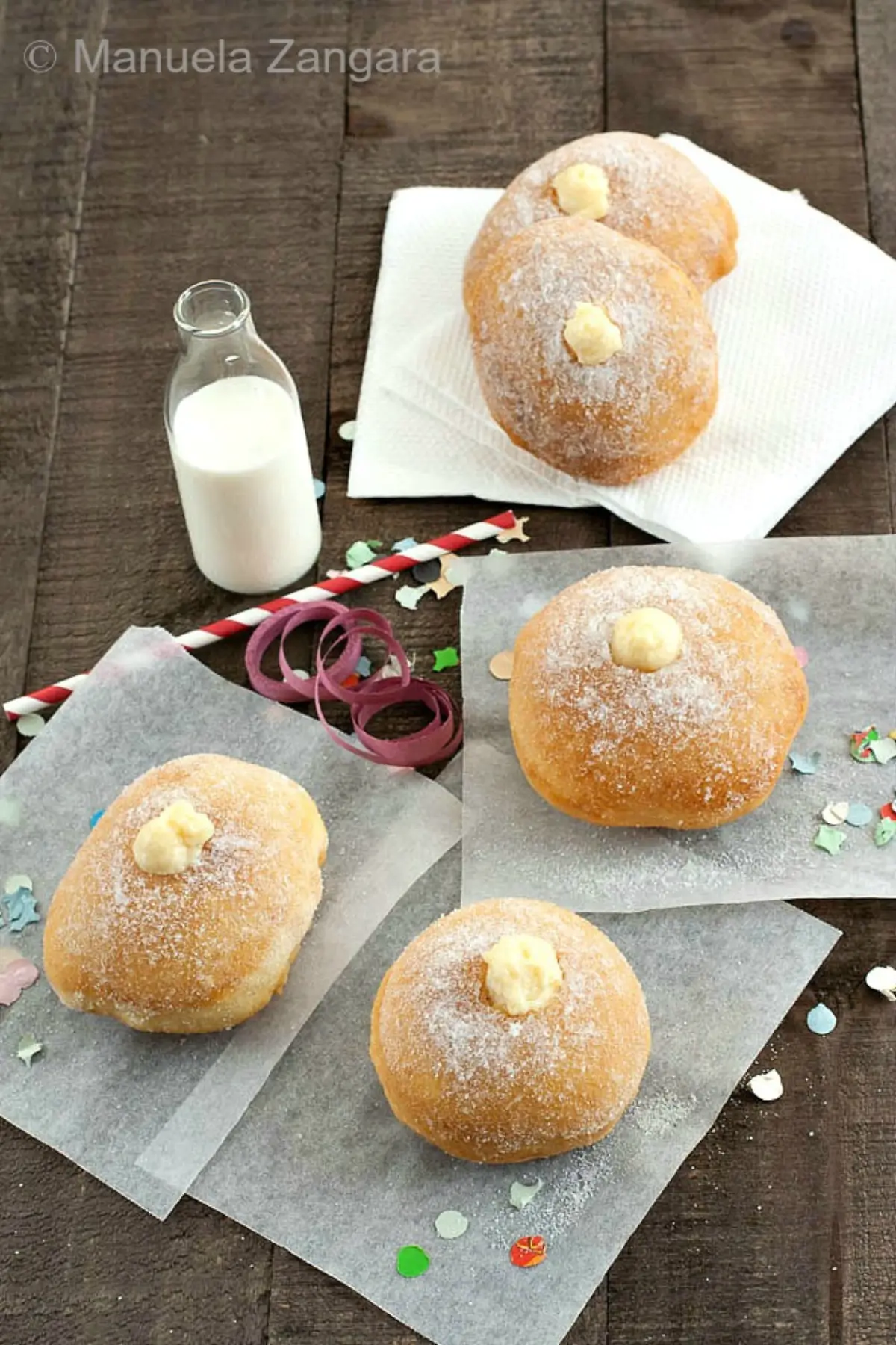 Overhead scene with sugar-dusted bomboloni filled with crema pasticcera, served with milk and Carnival confetti.