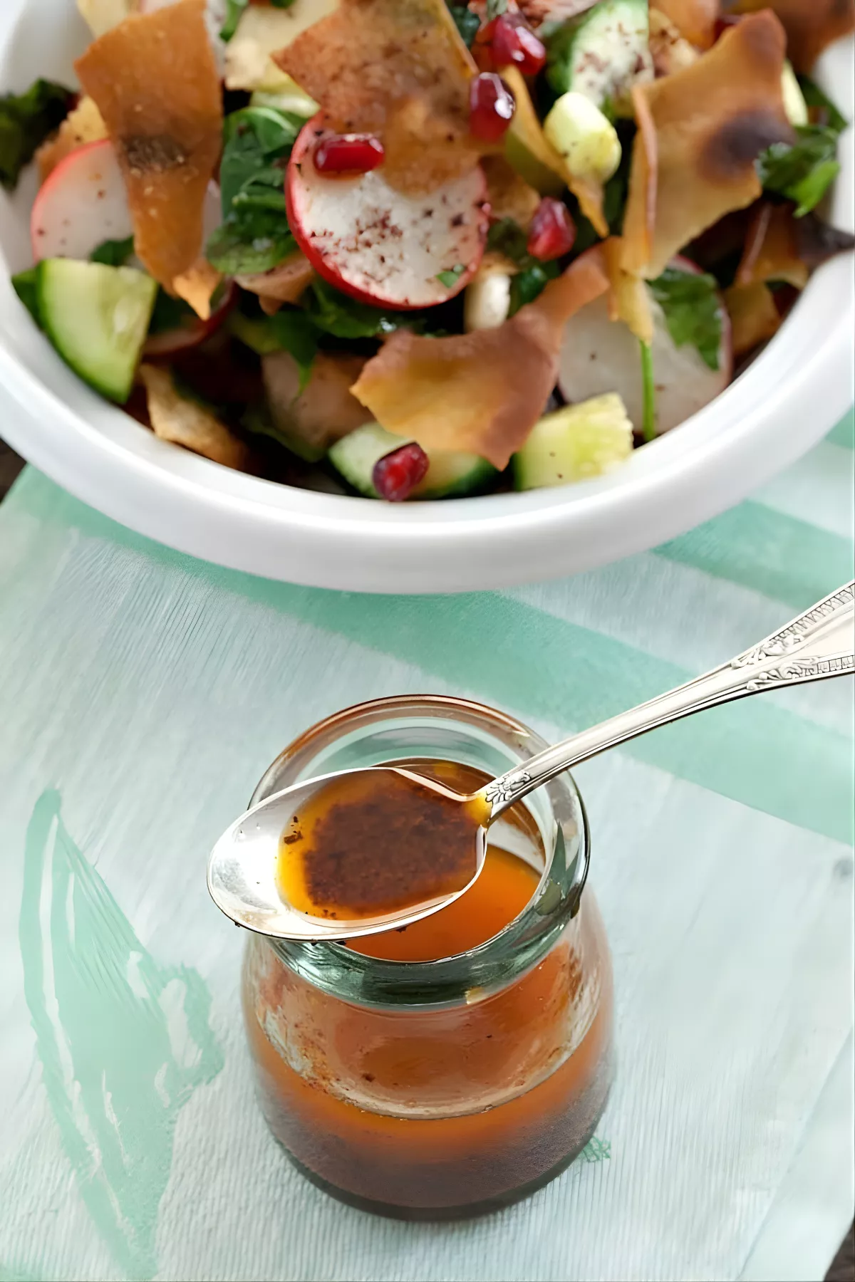 A spoonful of sumac dressing being poured from a jar over Fattoush salad.