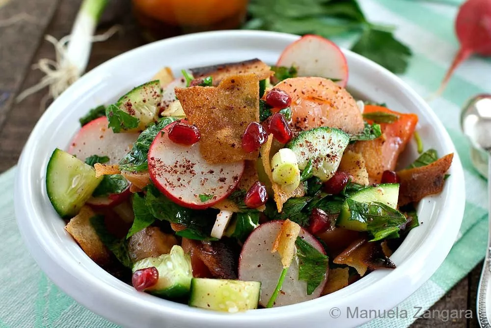 A vibrant bowl of Fattoush salad, topped with crispy khubz bread, cucumber, radish, pomegranate, and fresh herbs.