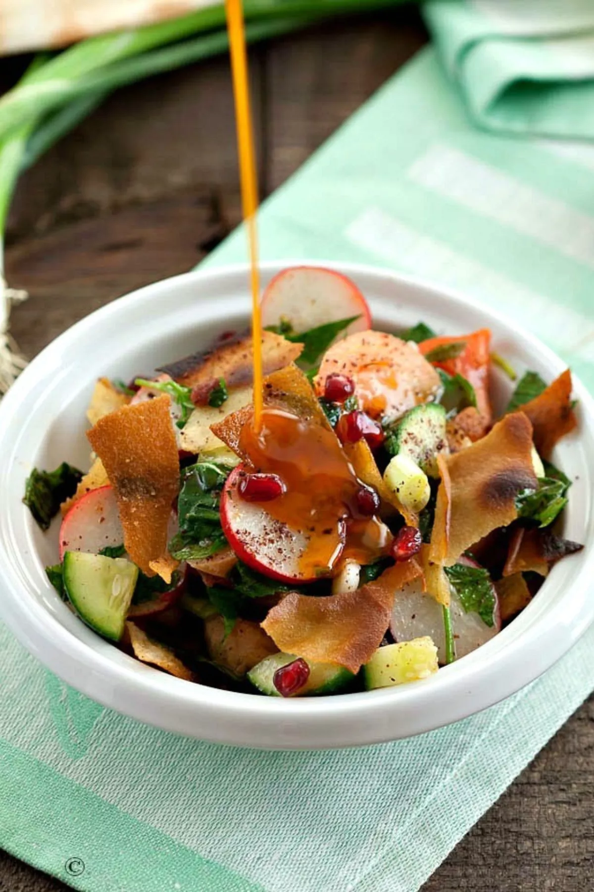 A spoonful of sumac dressing being poured over a bowl of Fattoush salad.