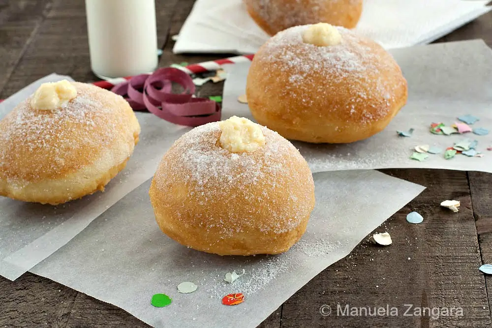Sugar-dusted bombolone topped with crema pasticcera on parchment paper.