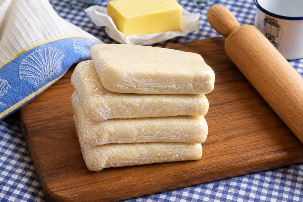 Wrapped puff pastry portions stacked on a wooden board beside butter and a rolling pin.