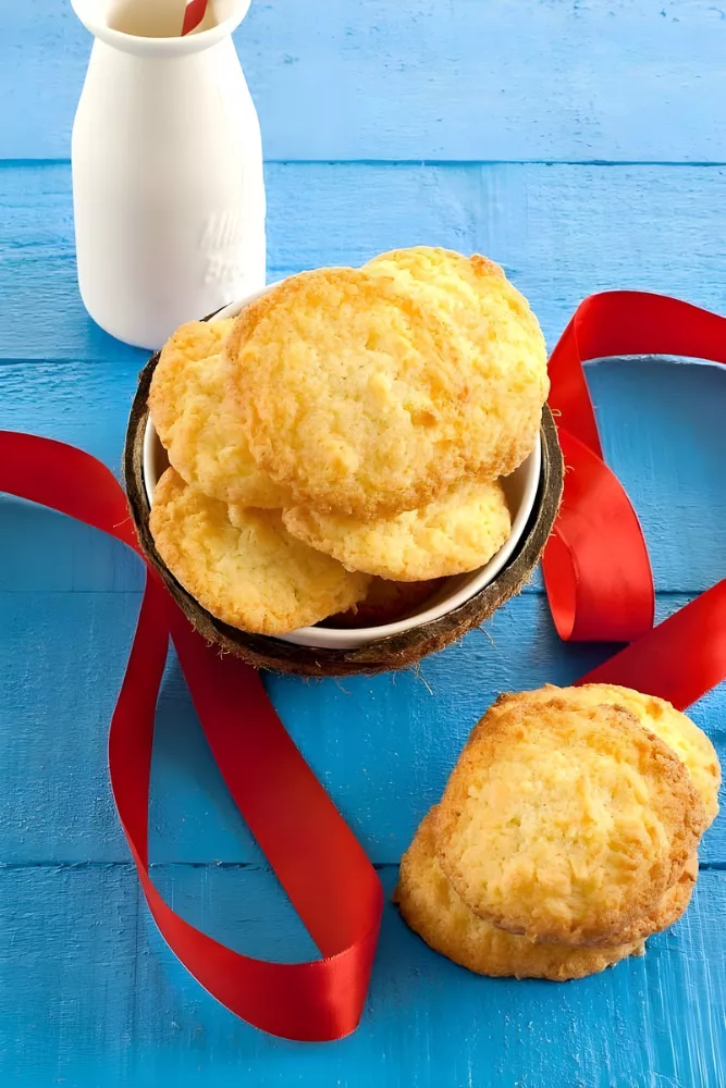 A bowl filled with golden coconut cookies with a red ribbon on a blue table.