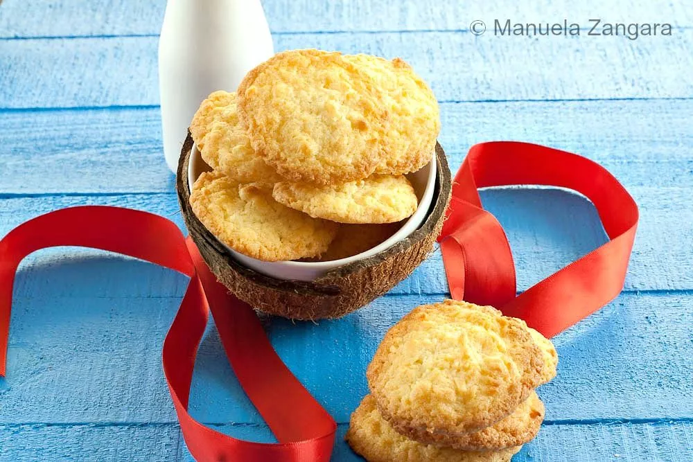 Coconut cookies served in a coconut bowl beside a bottle of milk on a blue background.
