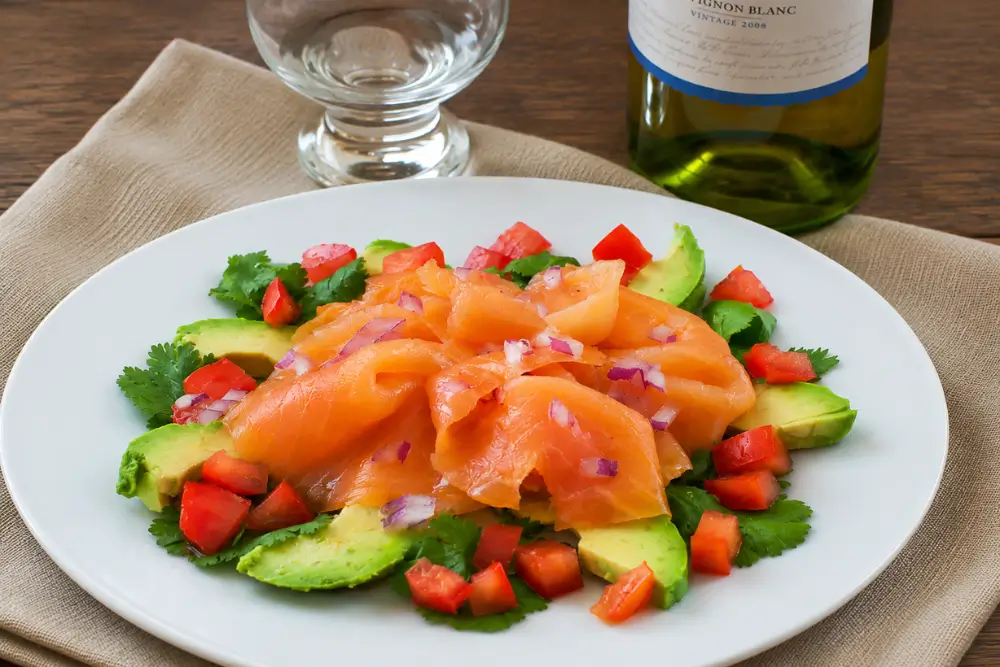 Smoked salmon served with sliced avocado, chopped tomato, cilantro, and red onion on a white plate.
