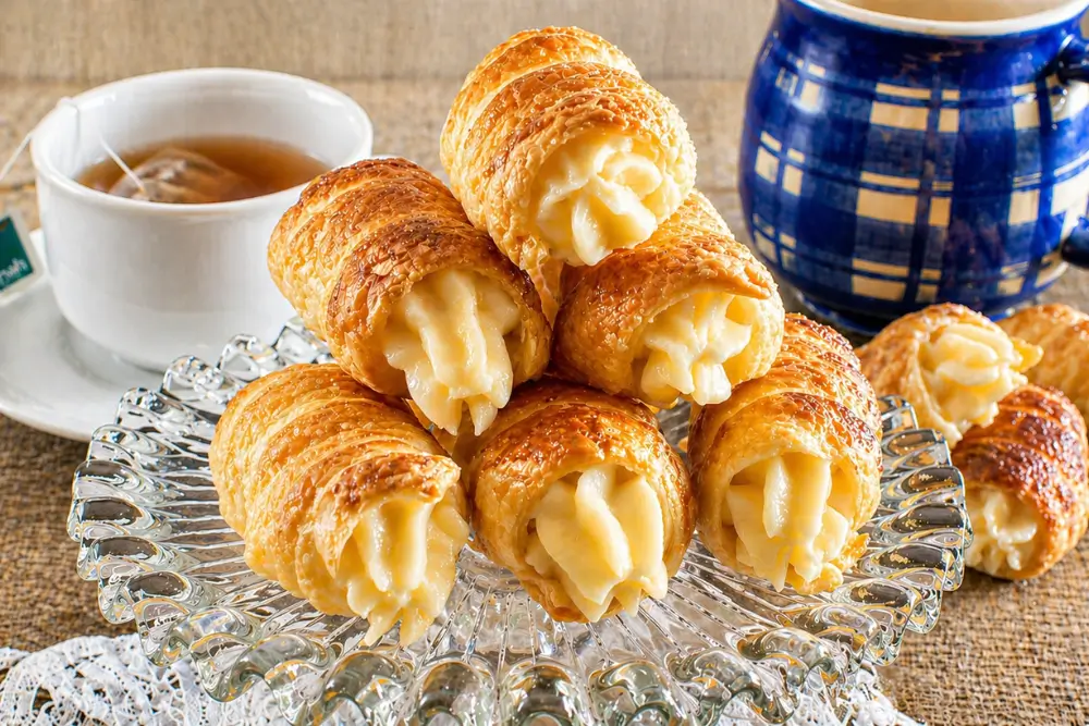 Cannoncini alla Crema filled with crème pâtissière served on a glass cake stand beside tea and coffee.