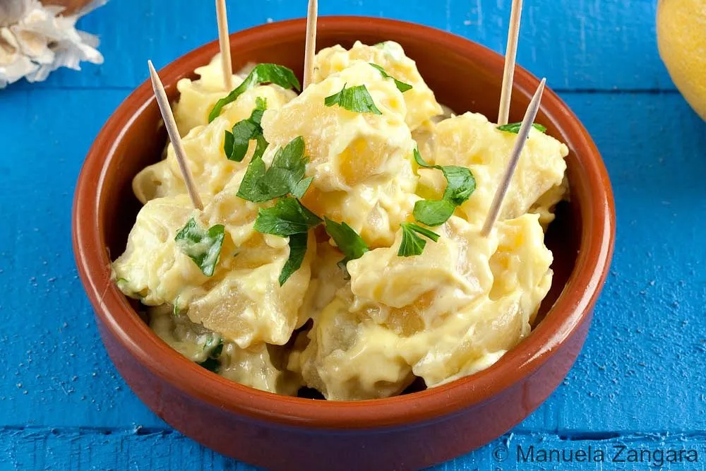 Creamy Patatas Alioli served in a bowl with aioli and garlic cloves nearby.