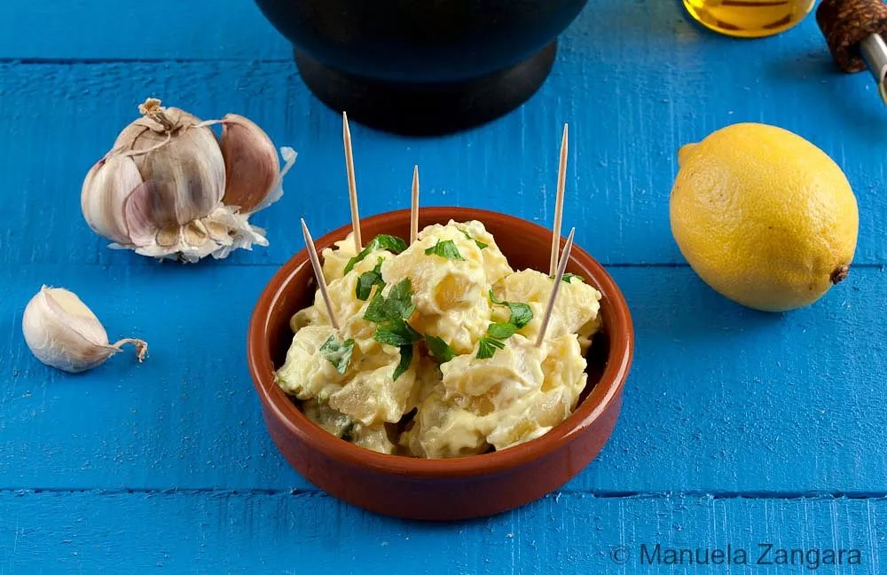 Spanish-style potatoes in aioli garnished with parsley, surrounded by garlic and lemon.