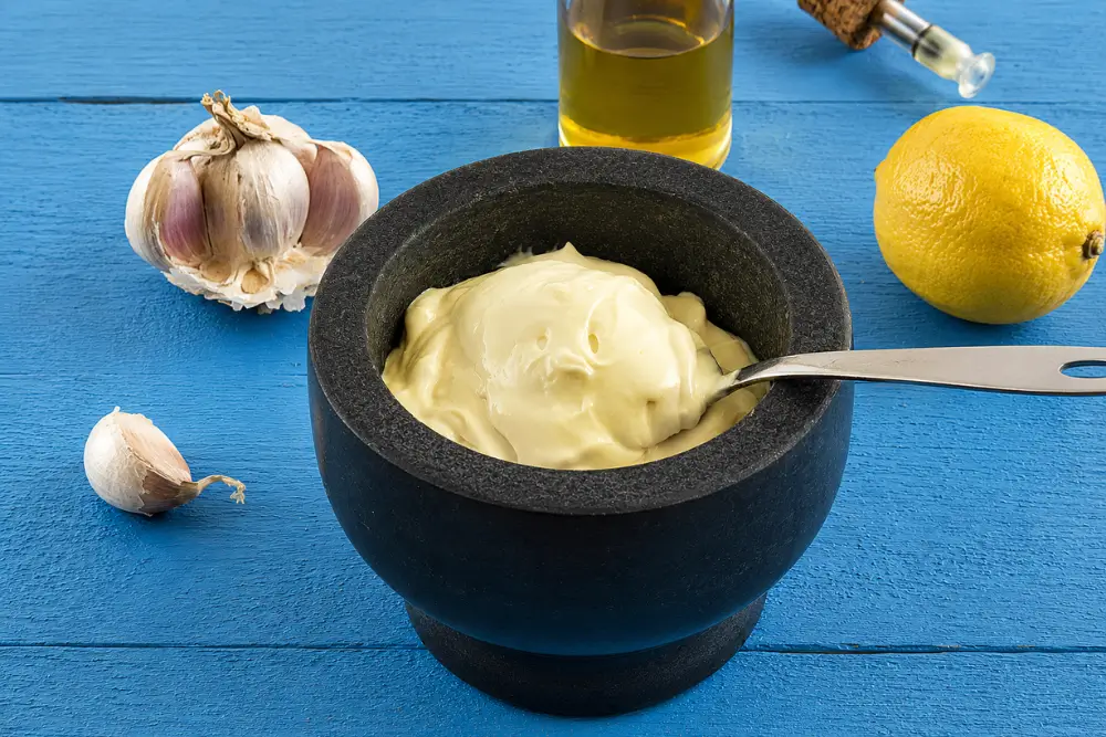 Spoon lifting thick and creamy homemade aioli from a black serving bowl.